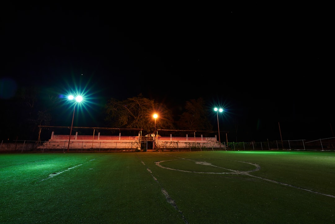 a soccer field at night with green lights friday night lights, high school football game, packed stadium cheering