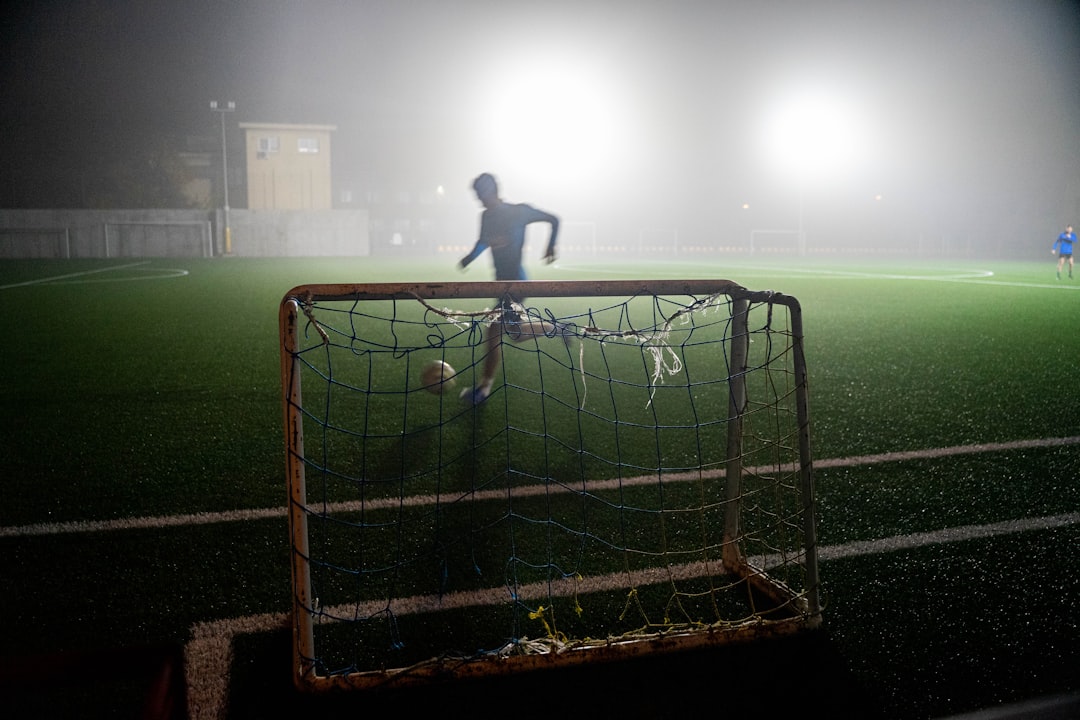 man in black shirt standing on soccer goal net during night time soccer player scoring goal close range stadium lights