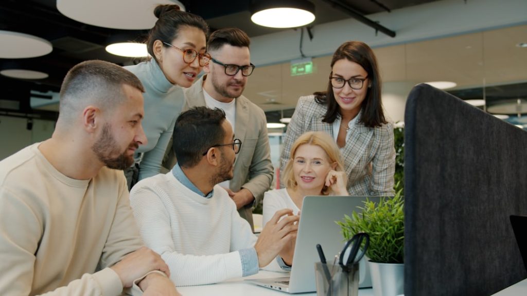 Diverse team collaborating around a laptop in office. team collaboration software dashboard, knowledge base interface, modern office workspace